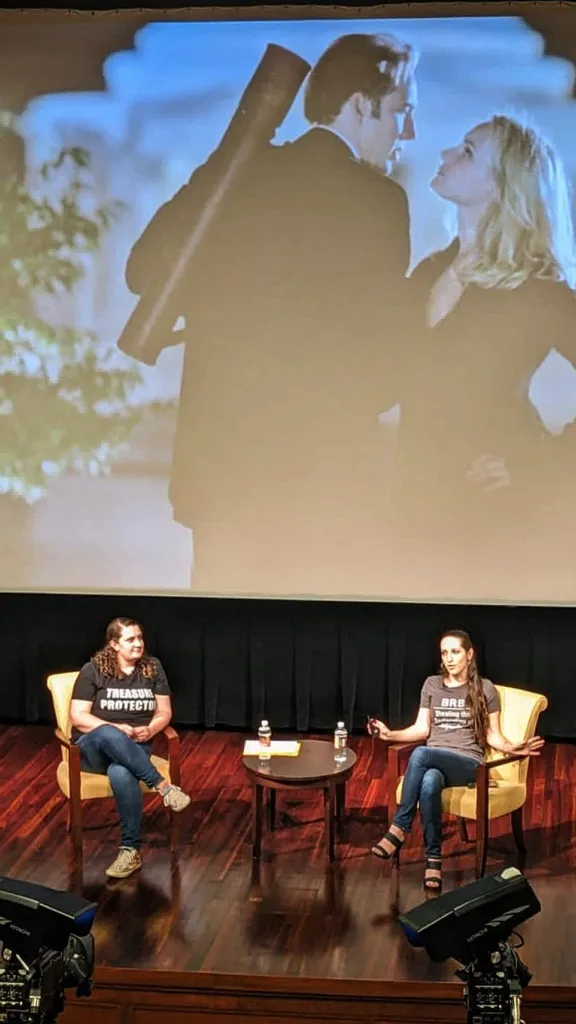 National Treasure Hunt Podcast hosts Aubrey Paris and Emily Black give a lecture on stage. A scene from National Treasure is projected on a screen behind them.